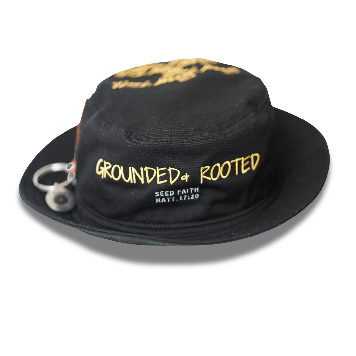Front close-up of the “Grounded & Rooted” black bucket hat by Cotton Faith, embroidered with gold lettering and the scripture “Seed Faith, Matt. 17:20,” with a chrome seed tag accessory on the brim.