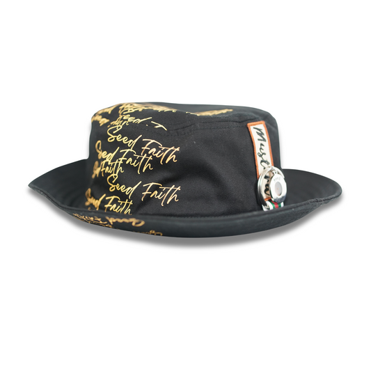 Back angle of the Cotton Faith “Grounded & Rooted” bucket hat showcasing all-over gold “Seed Faith” print, Mustard fabric label, and chrome key ring accessory on black cotton.