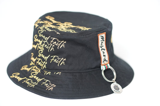 Close-up side view of the black “Grounded & Rooted” Mustard Seed Faith bucket hat by Cotton Faith, featuring gold “Seed Faith” script across the crown and a woven Mustard tag with attached chrome ring and pendant.